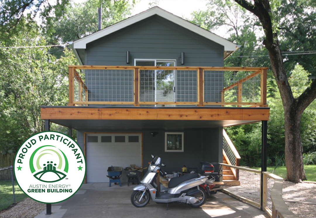 Two-story accessory dwelling unit in Austin, Texas with dark gray siding, cedar deck railing, and covered carport below, featuring an Austin Energy Green Building 'Proud Participant' badge