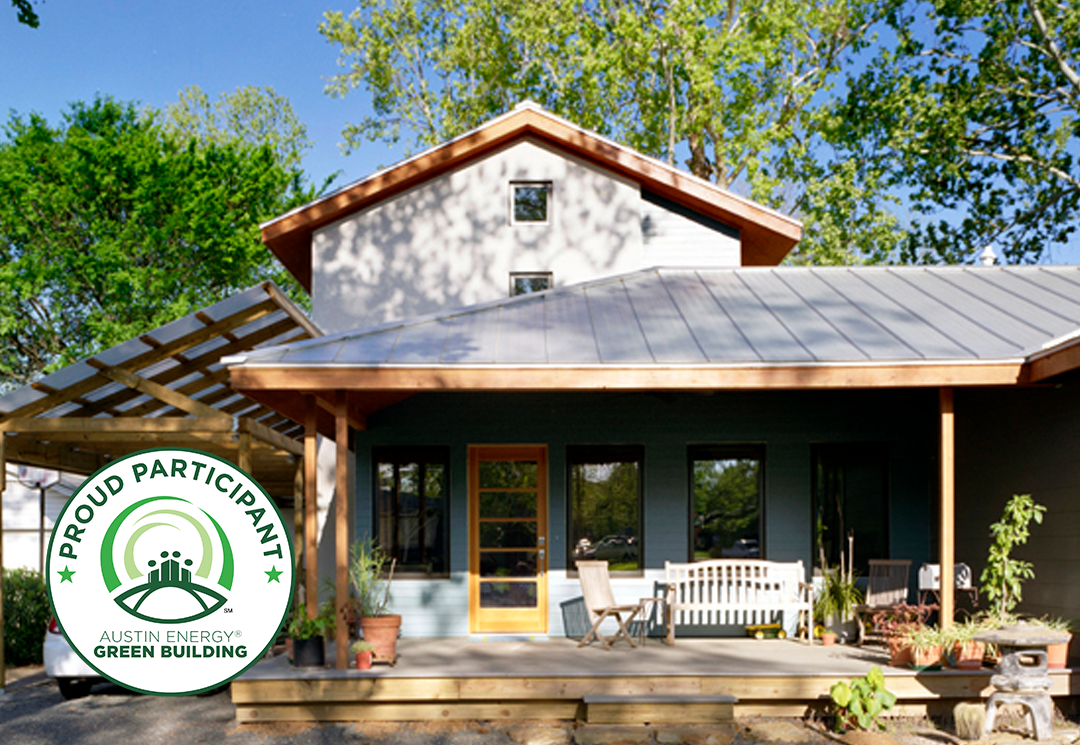 Renovated home exterior with dark green siding, metal roof, wood-trimmed gable, and covered porch; Austin Energy Green Building "Proud Participant" badge in corner.