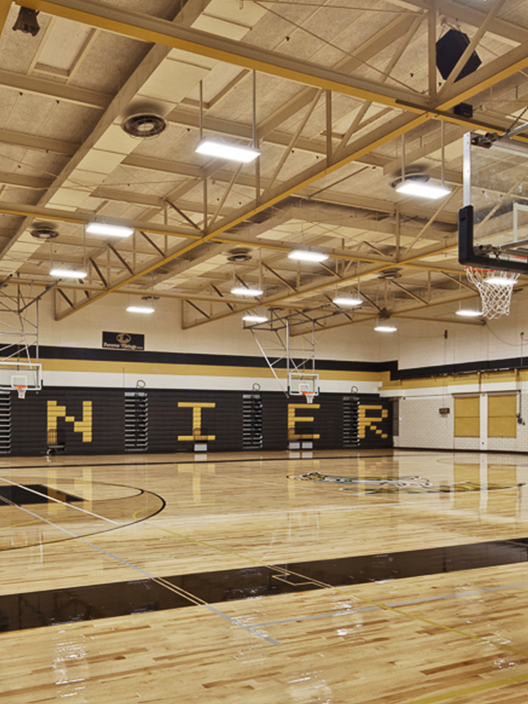 Interior of a school gymnasium at an Austin Independent School District facility, showing the hardwood court, bleachers, and overhead lighting