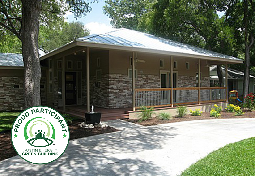 Exterior view of a house with stone veneer siding, a metal roof, and a covered front porch, surrounded by trees and landscaping. An "Austin Energy Green Building — Proud Participant" badge is overlaid in the bottom left corner.