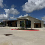 New single-story commercial building in Manor, Texas, with stone and dark metal facade, storefront glazing, and a freshly paved parking lot