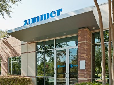Exterior entrance of the Zimmer Biologics facility featuring the company sign, brick columns, and glass facade