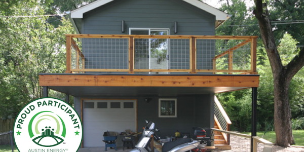 Two-story accessory dwelling unit in Austin, Texas with dark gray siding, cedar deck railing, and covered carport below, featuring an Austin Energy Green Building 'Proud Participant' badge