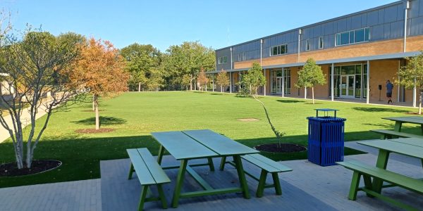 Outdoor patio and artificial turf at Casis Elementary School with green picnic tables, planters, and the school building in the background