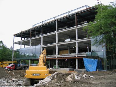 Steel-framed building under construction with a yellow excavator working at the base, photographed from ground level.