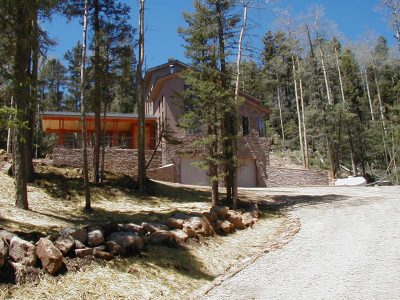 Exterior of a mountain home with stone and wood siding nestled among tall pine trees, with a gravel driveway and natural rock landscaping.