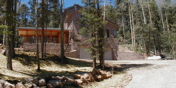 Exterior of a mountain home with stone and wood siding nestled among tall pine trees, with a gravel driveway and natural rock landscaping.