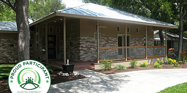 Exterior view of a house with stone veneer siding, a metal roof, and a covered front porch, surrounded by trees and landscaping. An "Austin Energy Green Building — Proud Participant" badge is overlaid in the bottom left corner.