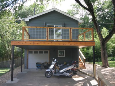 Rear exterior of a two-story urban home featuring a rooftop deck with wood and cable railing, garage below, and mature trees surrounding the property.